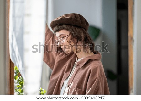 Pensive melancholic teenager in vintage clothes, beret and glasses standing at window ang looking attentively. Teen girl with concentrated glance looks like dreamer, bookworm, brainy diligent student  Royalty-Free Stock Photo #2385493917
