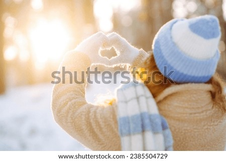 A woman in a hat and scarf against the backdrop of a snowy winter forest holds her hands in the shape of a heart. Back view. Young woman enjoying a sunny winter day. Vacation concept, lifestyle.