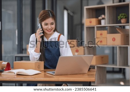 A portrait of a young Asian woman, e-commerce employee sitting in the office full of packages in the background write note of orders and use smartphone, for SME business ecommerce and delivery.