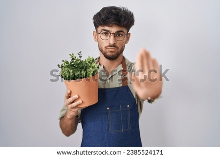 Arab man with beard holding green plant pot doing stop sing with palm of the hand. warning expression with negative and serious gesture on the face. 