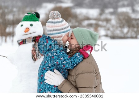 Portrait of happy mother and child hugging in winter outside. Child embracing mom against the snowman wearing hat and scarf