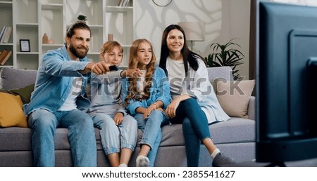 Happy man and woman relaxing with daughter and son watching television at home together. Cheerful family relaxing on sofa at home watching movie with children.