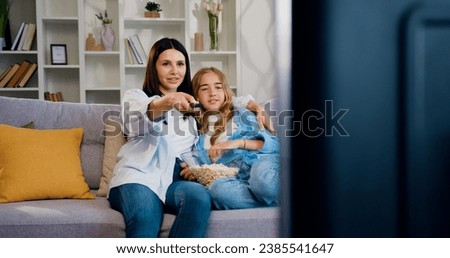 Beautiful caucasian mother and douther spend time sitting on sofa at home, eating popcorn and watching TV. Family enjoy funny television show, smiling mother holding remote control watching cartoons