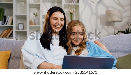 Close up portrait of happy mother with her daughter using laptop at home in living room. Young woman watching cartoons on laptop with girl and smiling.