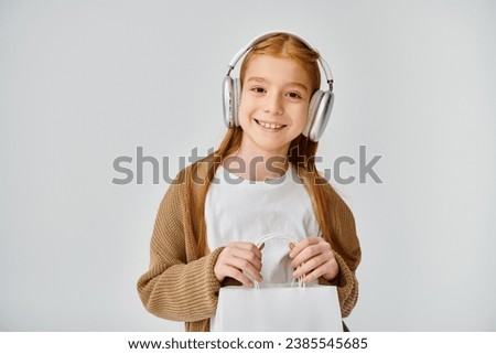 happy preteen girl in casual outfit with headset holding present bag and smiling at camera, fashion Royalty-Free Stock Photo #2385545685