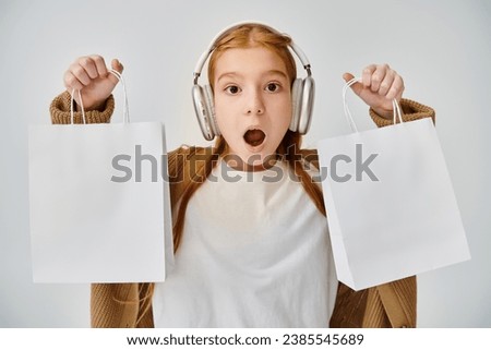 astonished little girl with headphones showing present bags at camera, Black Friday, fashion concept Royalty-Free Stock Photo #2385545689