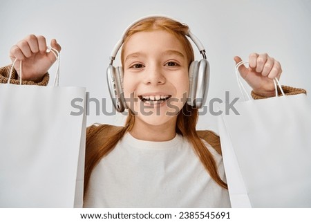 joyous little girl with modern headset posing with present bags on gray backdrop, fashion concept Royalty-Free Stock Photo #2385545691