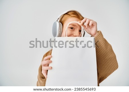 cheerful little girl in warm attire with headset and with present bag in front of her face, fashion Royalty-Free Stock Photo #2385545693