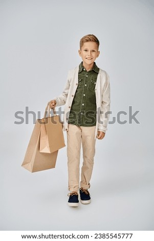 joyous little boy in stylish casual cardigan posing on gray backdrop with present bags, fashion Royalty-Free Stock Photo #2385545777