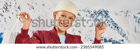 preteen boy in stylish clothes holding present bags and smiling at camera, fashion concept, banner Royalty-Free Stock Photo #2385549085