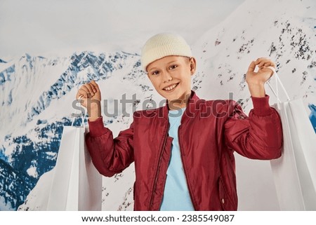 little boy in stylish clothes and beanie hat holding present bags and smiling at camera, fashion Royalty-Free Stock Photo #2385549087