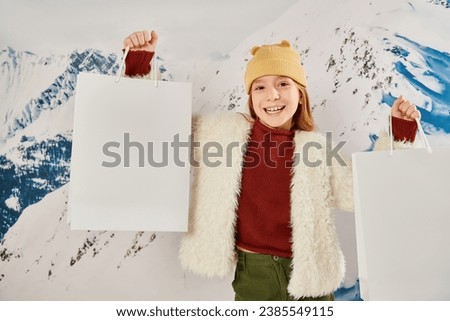 joyous girl in beanie hat and winter jacket holding present bags and smiling at camera, fashion Royalty-Free Stock Photo #2385549115