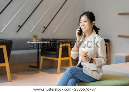 Cheerful Asian woman uses smartphone to work while drinking coffee in living room at home.