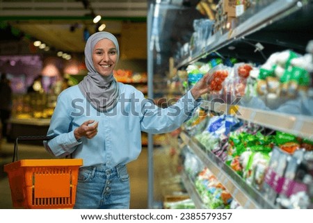 Portrait female shopper, Muslim woman in hijab smiling and looking at camera, choosing vegetables and fruits in a large supermarket store, holding a basket with goods in hands, grocery department. Royalty-Free Stock Photo #2385579357