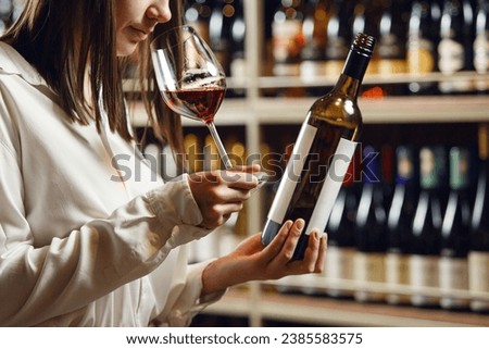 Professional woman sommelier smells red wine from glass, standing in cellar against shelf various alcoholic beverage background. Royalty-Free Stock Photo #2385583575