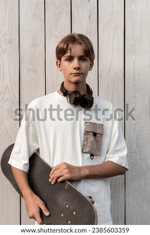 Teen boy with headphones holding skateboard outdoor on wooden white background. Child spending free time relaxing, walking. Technology, teen hobby, active lifestyle and people concept.