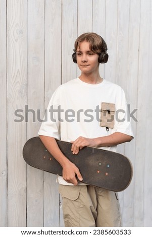 Teen boy in headphones holding skateboard outdoor on wooden white background. Child spending free time relaxing, walking, listen music. Technology, teen hobby, active lifestyle and people concept.