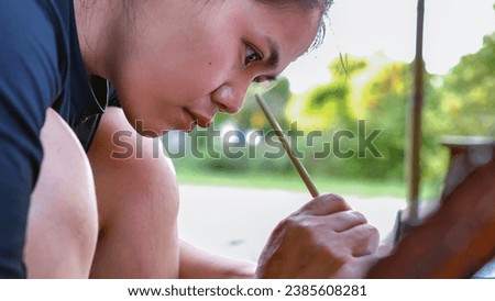 Close-up of Asian woman painter creating art use a paintbrush to draw lettering designs on a wooden coffee shop sign. outdoor activities, People doing activities.