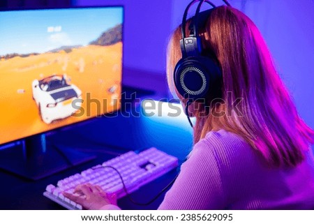 Happy Gamer endeavor plays online video games tournament with computer with neon lights, Young player woman wearing gaming headphones intend to do playing car games online at home, Back view Royalty-Free Stock Photo #2385629095