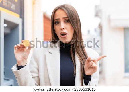 Young pretty woman holding a Bitcoin at outdoors surprised and pointing side