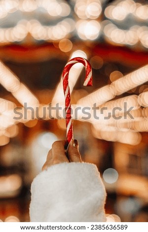 Female hand holding red white cane sweet lollipop against the background of golden fairy lights. Christmas holiday magic details