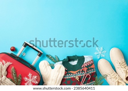 Yuletide voyage concept. Top view picture showcasing red suitcase, winter boots, cozy scarf, sweater, mittens, pine twigs, snowflakes, baubles on vibrant blue surface, perfect for text or advertising