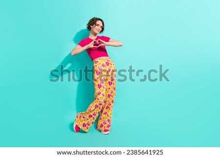 Full length photo of cheerful cute woman dressed flower print trousers showing arms heart empty space isolated teal color background