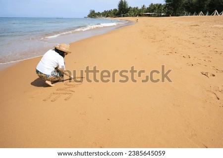 A woman sits on the sand and wearing a hat writing the letters L O V E in the sand on a holiday morning. A young female tourist happily draws "love" on the sand at the beach while relaxing. 
 Royalty-Free Stock Photo #2385645059