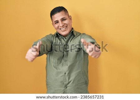 Hispanic young man standing over yellow background approving doing positive gesture with hand, thumbs up smiling and happy for success. winner gesture.  Royalty-Free Stock Photo #2385657231