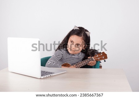 Picture of girl learning to play ukulele on a laptop with light background. Concept of music and learning.
