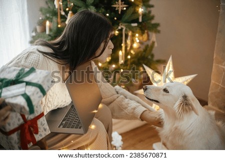 Happy woman holding stylish christmas gifts with credit card and caressing cute white dog in festive decorated christmas room with lights. Christmas shopping online and black friday sales.