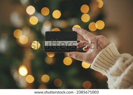 Hand in cozy sweater holding credit card close up on background of christmas tree lights in festive decorated room. Christmas shopping online and black friday sales.