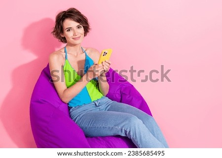 Photo of beautiful cute young girl sitting beanbag browsing her brand new iphone 15 after release isolated on pink color background