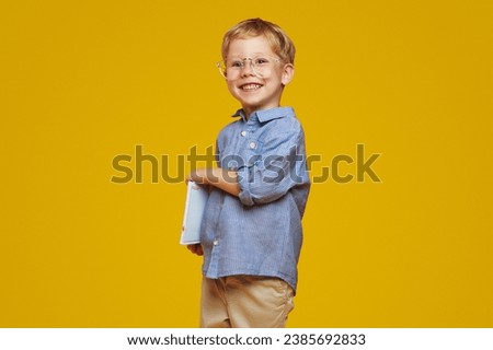 Happy little boy wearing nerdy glasses and blue shirt holding notepads and smiling at camera while standing against yellow background.