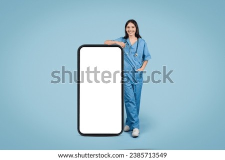 Happy young female healthcare worker in scrubs standing near huge cellphone with empty screen, ready for medical custom text, full length, mockup