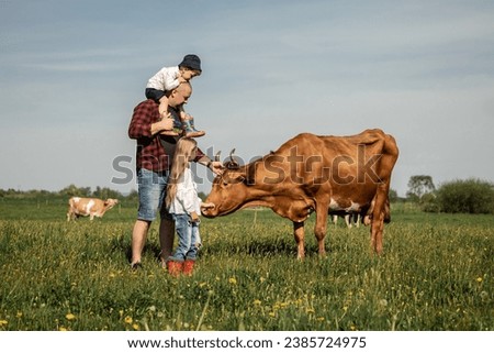 Happy farmers family in green field with big cow in a green field with flowers on a sunny summer day. Royalty-Free Stock Photo #2385724975