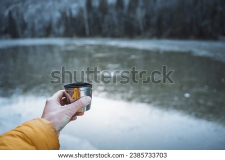 Hand with mug on the background of a frozen lake, travel photo, mug with hot drink in nature. A metal tumbler in a man's hand. High quality photo