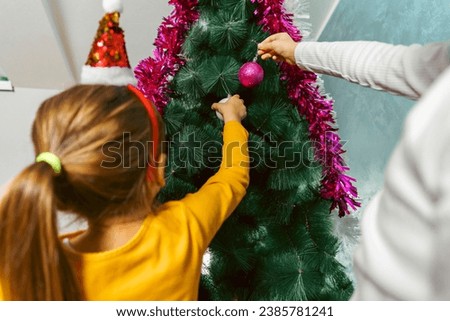Picture of a girl decorating christmas tree in a living rom, wearing fun festive headband. Mother's arm in the frame putting ornaments on the tree