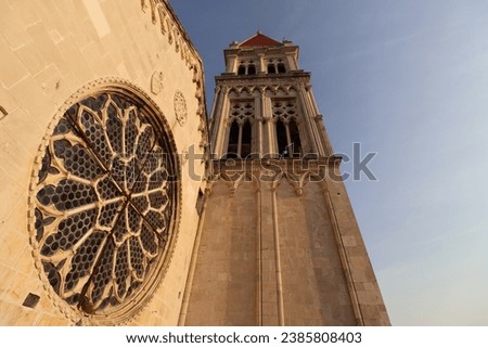 Wode angle photography towards top of a bell towe in Trogir, Croatia. Side wall of the church with big rosetta. Sandstone in sunset lightning.  Royalty-Free Stock Photo #2385808403