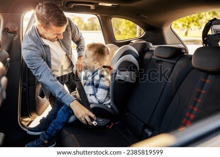 A cheerful dad is buckling up his son in his car seat and preparing for trip. Royalty-Free Stock Photo #2385819599