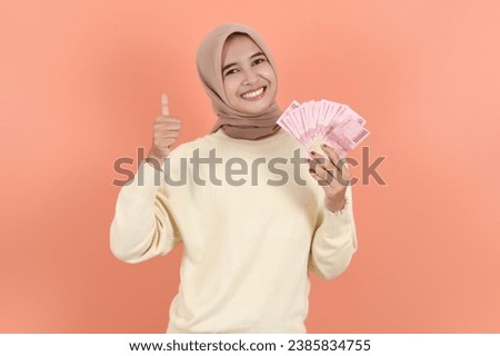 Smiling beautiful Asian Muslim woman in cream sweater holding cash money in Indonesian rupiah banknotes, gesturing thumb up  isolated over peach background. ​People religious lifestyle concept Royalty-Free Stock Photo #2385834755