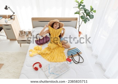 Top view shot of Asian young happy female traveler wears big hat laughing smiling look at camera sitting trying on yellow summer long dress on bed preparing personal stuff for dream destination trip.