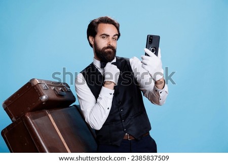 Elegant hotel employee takes photos while being on studio camera, posing with confidence and being dressed in formal hotel attire. Professional stylish bellhop using smartphone for pictures.
