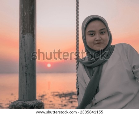 portrait of a teenage girl wearing a hijab sitting on a swing against a sunset background