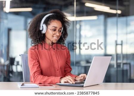 Young beautiful woman inside the office works with a laptop, a businesswoman in headphones listens music, podcasts, audio books and training course. Worker smiling , with curly hair and red shirt. Royalty-Free Stock Photo #2385877845