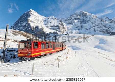 On a sunny winter day, tourists ride on a cogwheel train from Jungfraujoch (Top of Europe) to Kleine Scheidegg on the snowy hillside with Eiger and Monch in background, in Berner Oberland, Switzerland