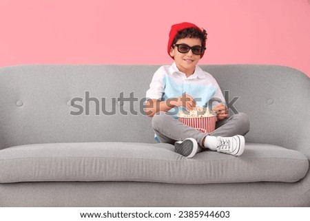 Little boy in 3D glasses with popcorn watching movie on sofa against pink background