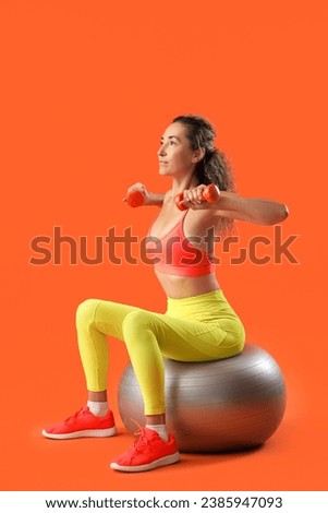 Beautiful adult woman exercising with dumbbells on fitness ball against orange background