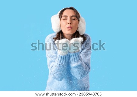 Young woman in warm clothes blowing snow on blue background