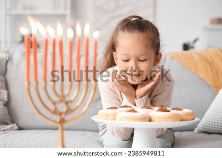 Little girl with donuts celebrating Hanukkah with menorah at home Royalty-Free Stock Photo #2385949811